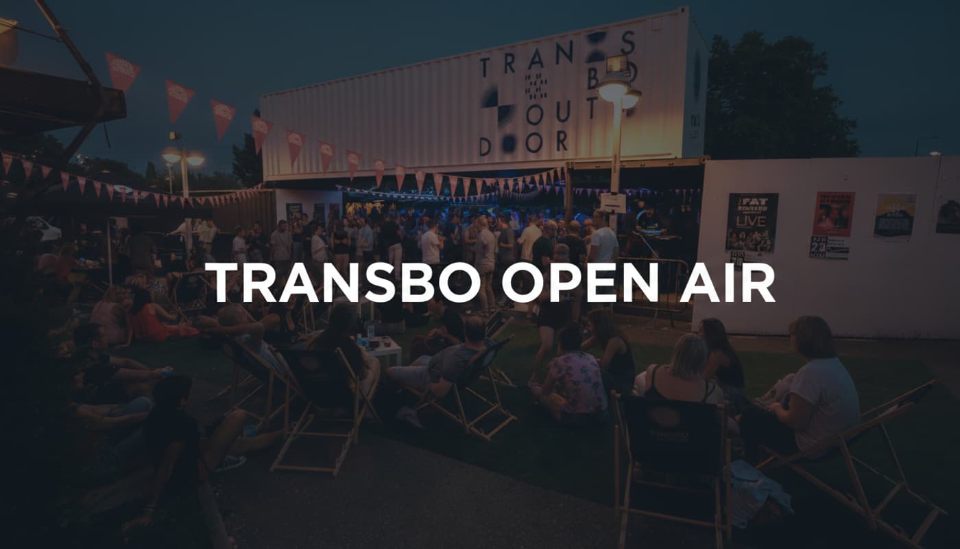 DJ mixant devant une foule sur la terrasse nocturne du Transbordeur sous des guirlandes d'ampoules avec l'inscription Transbo Open Air en surimpression.