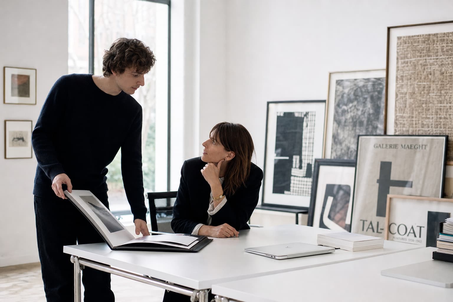 Un artiste présente son portfolio à un directeur de galerie assis devant un bureau blanc minimaliste dans une pièce lumineuse avec des cadres appuyés contre le mur.