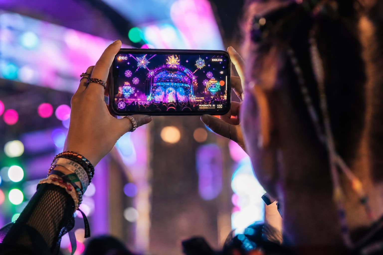 Un participant à un festival filme une scène avec son smartphone affichant un filtre de réalité augmentée coloré, avec des lumières néon en arrière-plan.