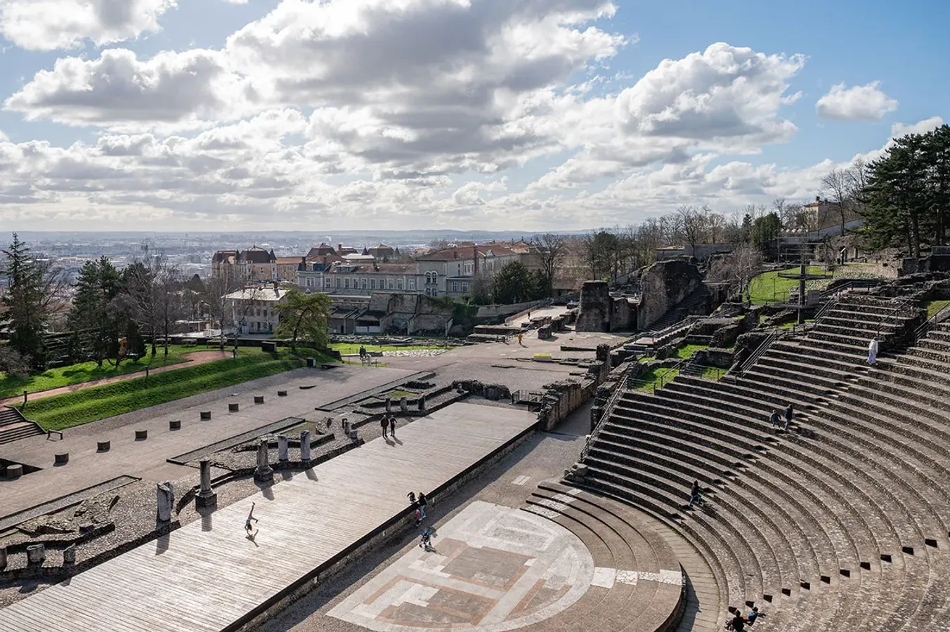 Vue panoramique du théâtre romain antique de Lugdunum sur la colline de Fourvière, avec ses gradins en pierre et les vestiges de la scène, la ville de Lyon visible en arrière-plan sous un ciel bleu.